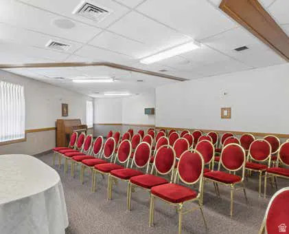 Spacious carpeted room featuring a drop ceiling with integrated lighting, wood trim accents, and a window with vertical blinds.