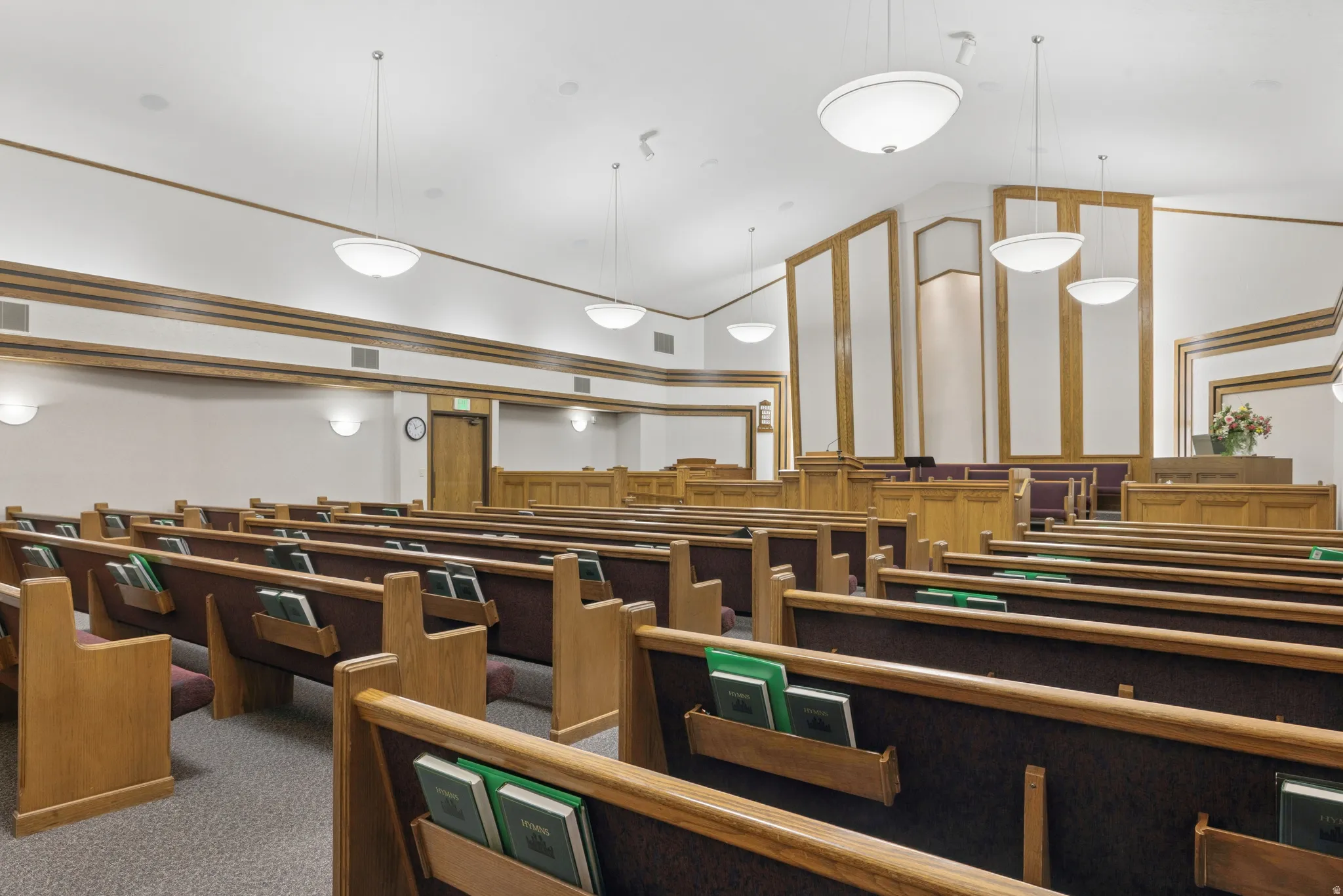 Spacious interior featuring multiple rows of built-in wooden seating with upholstered backs, integrated lighting, recessed wall panels with wood trim, and a high vaulted ceiling with pendant and wall sconce lighting.