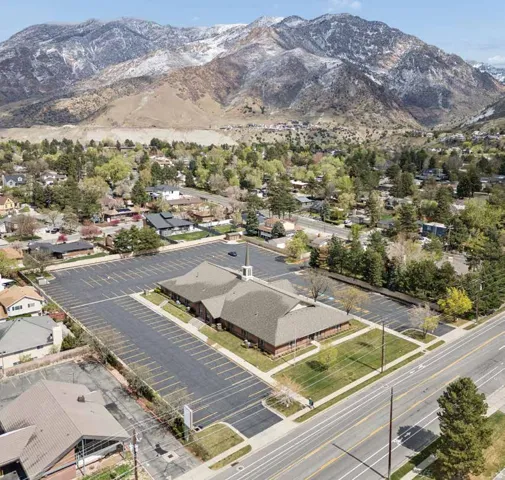 Spacious property featuring a large asphalt parking lot with marked spaces, a building with a gray shingle roof and brick siding, and surrounding green landscaping.
