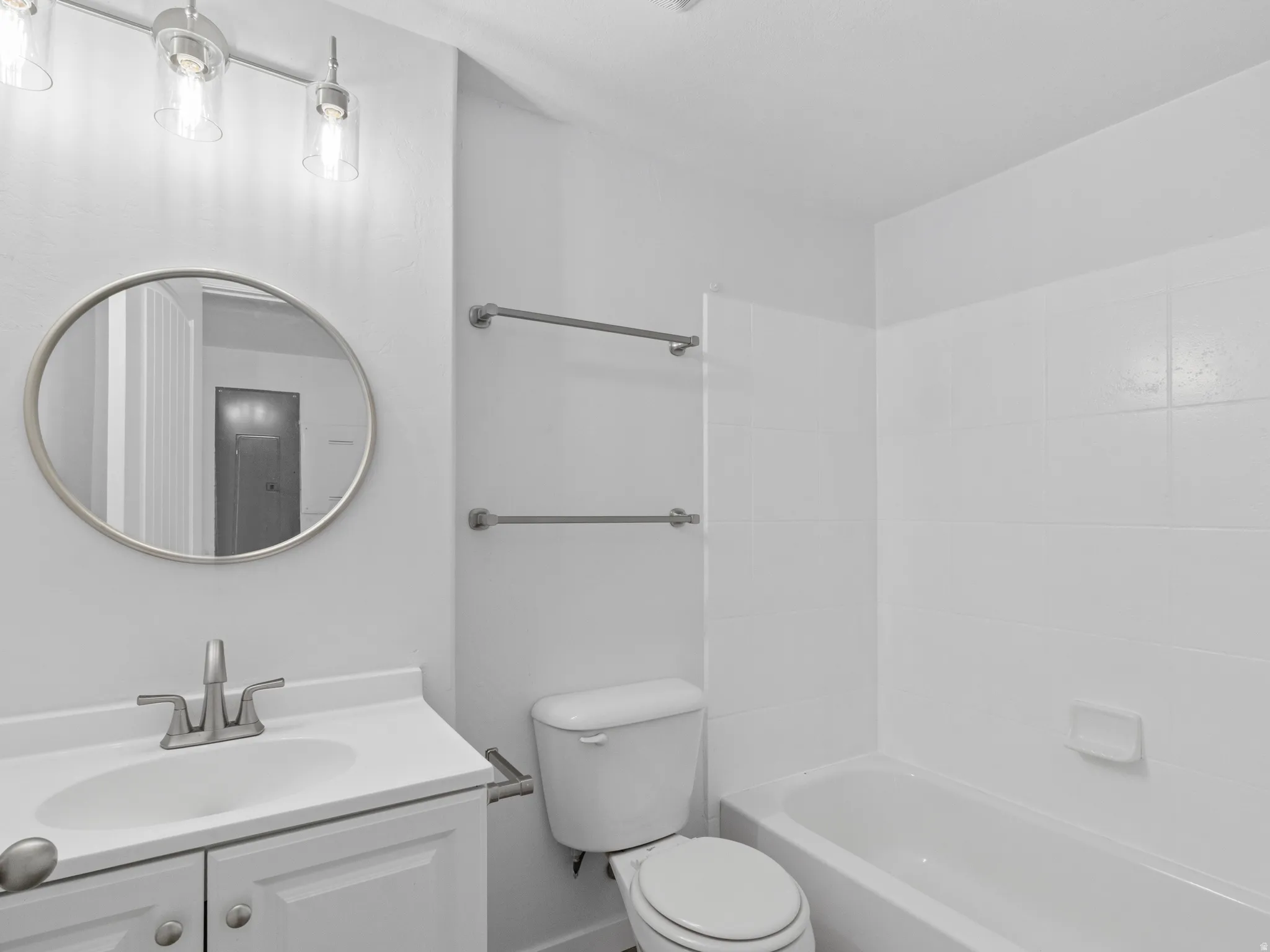 Bathroom featuring a single vanity with storage, a circular mirror, a track light fixture with clear glass shades, a toilet, and a bathtub with white tiled surround