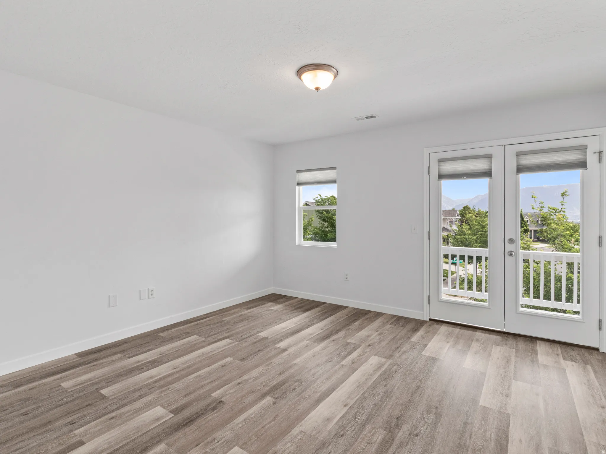 Double French doors opening to an outdoor balcony, complemented by wood-finish flooring, a contemporary ceiling light fixture, and a window providing natural illumination