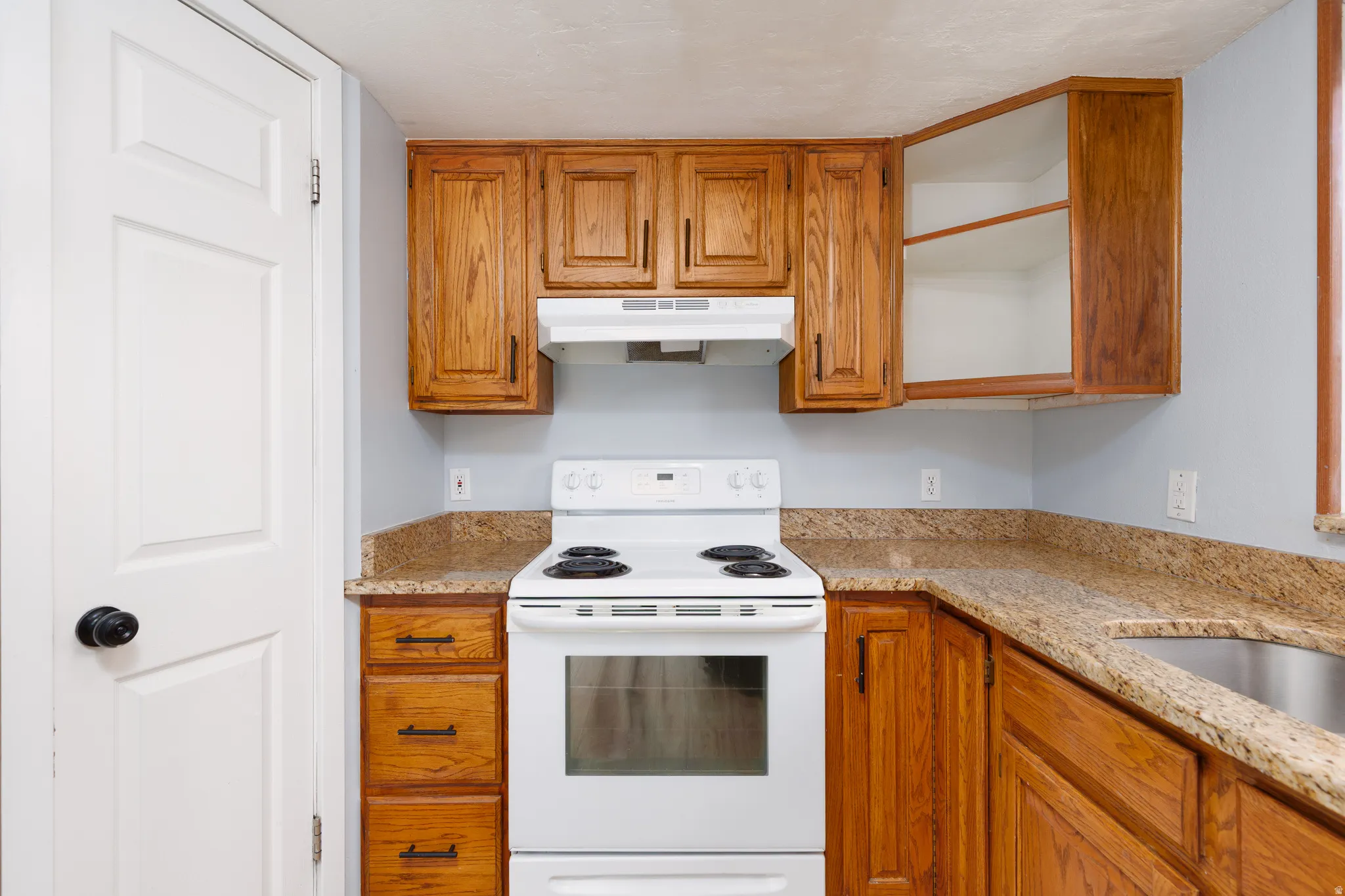Compact kitchen featuring wood cabinetry with black hardware, a white electric range, a matching range hood, and speckled countertops