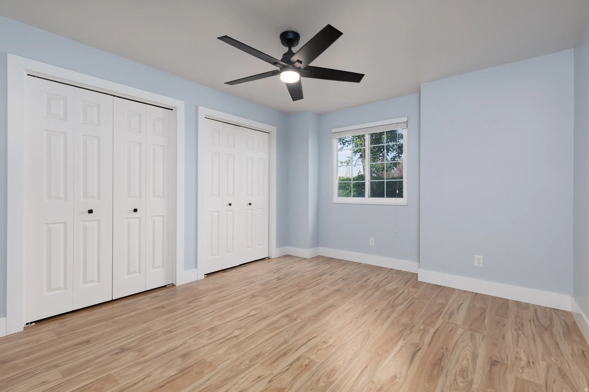 Open room featuring wood-finish flooring, two sets of bi-fold closet doors with black hardware, a ceiling fan with integrated lighting, and a window with white blinds