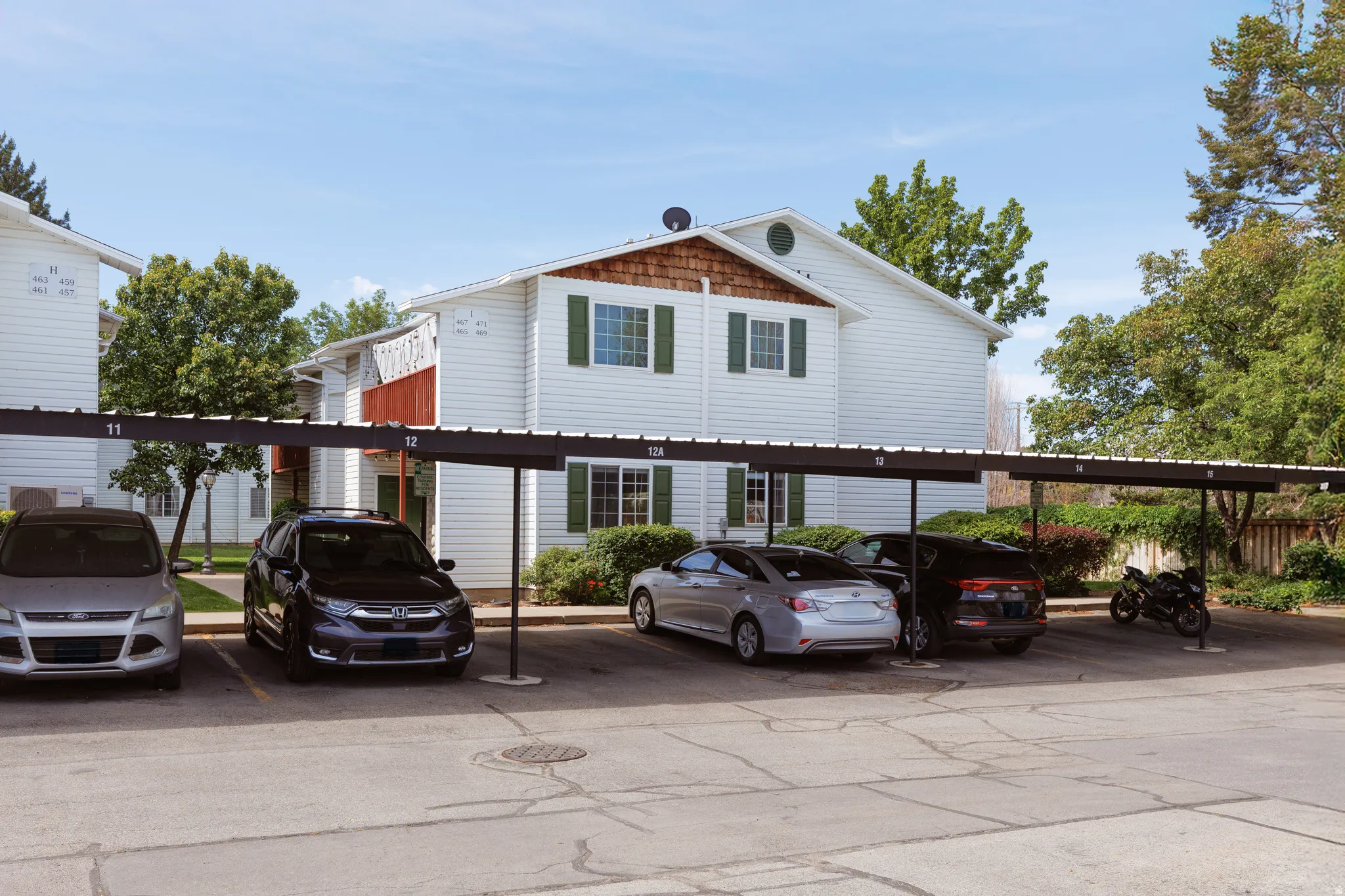Covered parking structure with individual numbered spaces, set against a two-story building featuring white horizontal siding, green window shutters, and a prominent round window