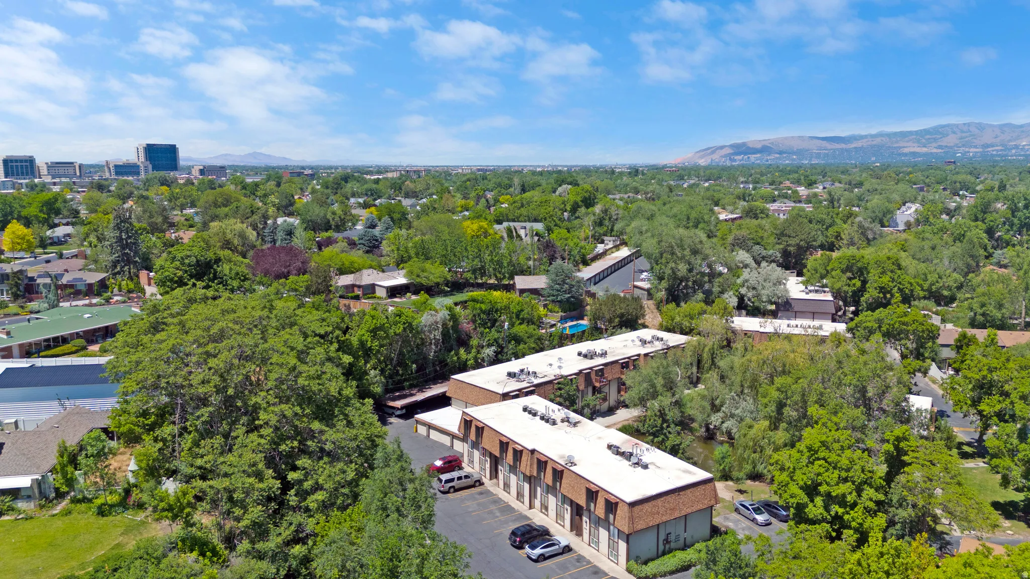 Aerial perspective showcasing multiple townhouse buildings with pitched roofs, surrounding lush green trees and a paved parking area