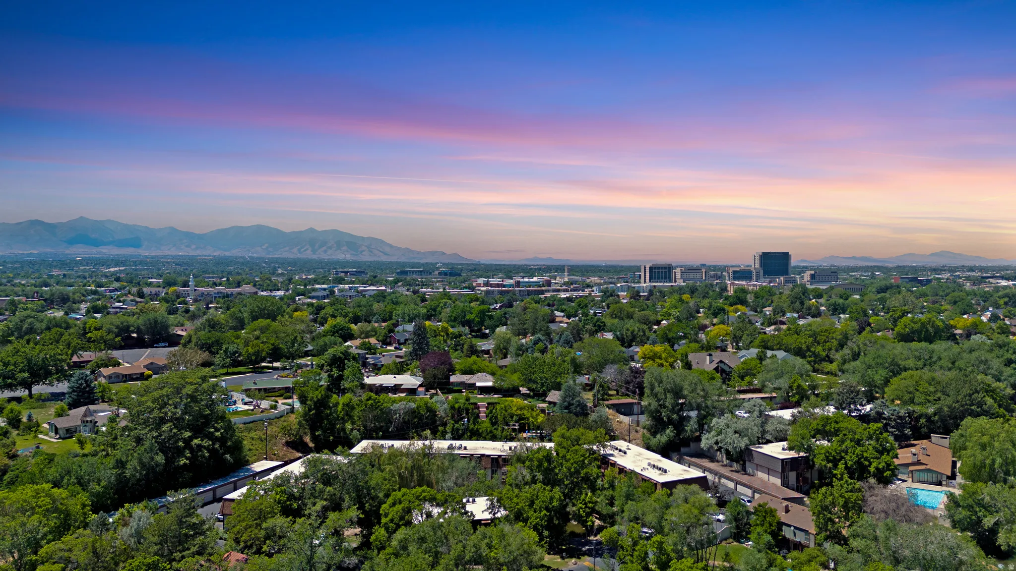 Expansive aerial cityscape featuring a dense canopy of mature trees, residential properties, distant mountain ranges, and commercial buildings