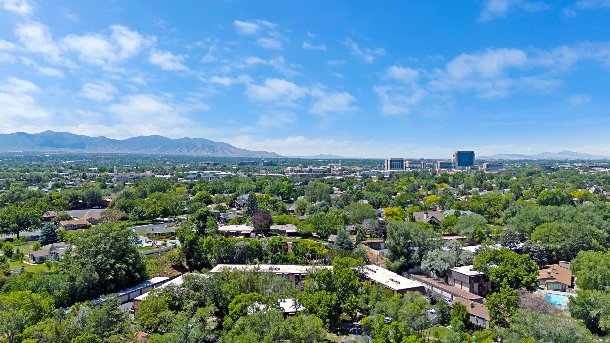 Expansive aerial city panorama featuring lush green trees, residential homes, distant mountains, and commercial buildings