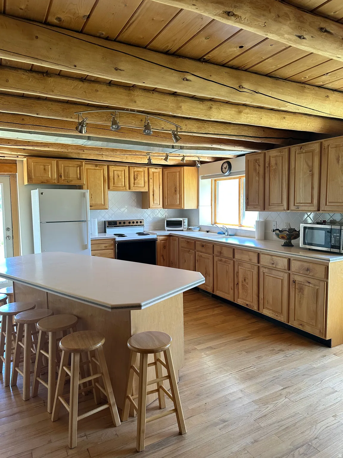 Spacious kitchen featuring light wood cabinetry, light countertops, wood-finish flooring, a central island with bar seating, and exposed wood ceiling beams