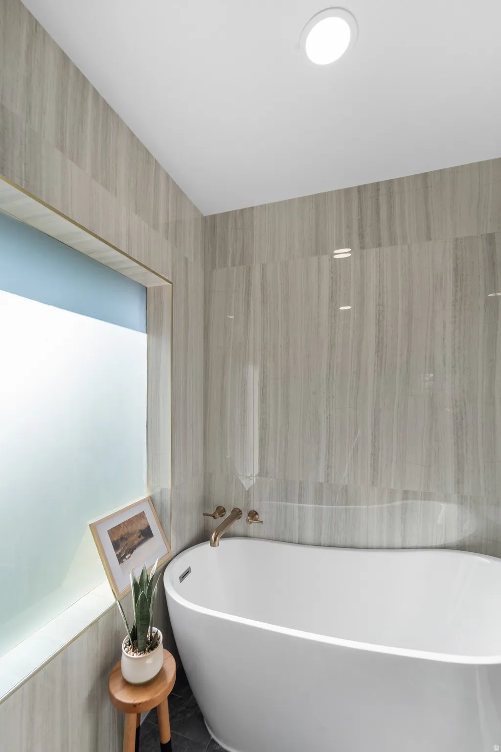 Freestanding soaking tub with wall-mounted brass-finish faucet, surrounded by large format stone-look wall tiles