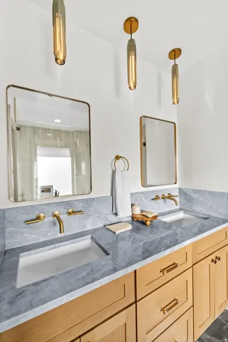Dual vanity featuring light-toned wood cabinetry with gold hardware, a grey veined countertop, and integrated rectangular sinks