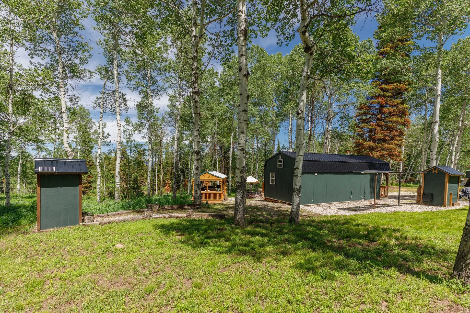 Expansive wooded setting featuring multiple outbuildings, including a primary green-sided structure with a black metal roof, a smaller green-sided structure, and a wooden gazebo