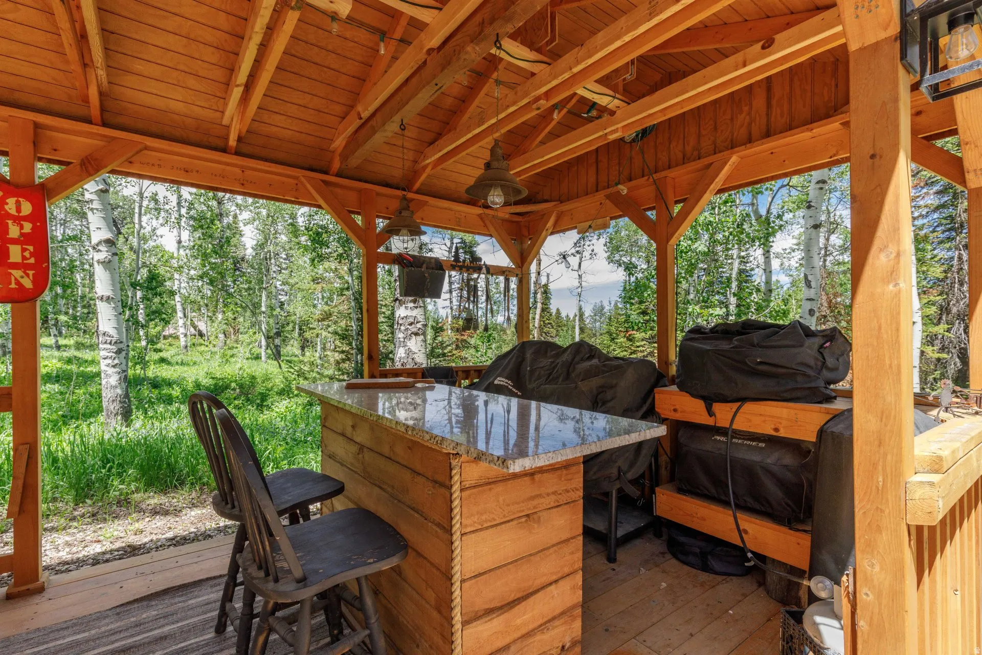 Open-air wood-framed gazebo featuring a built-in bar with a stone-finish countertop, wood flooring, and exposed timber ceiling beams