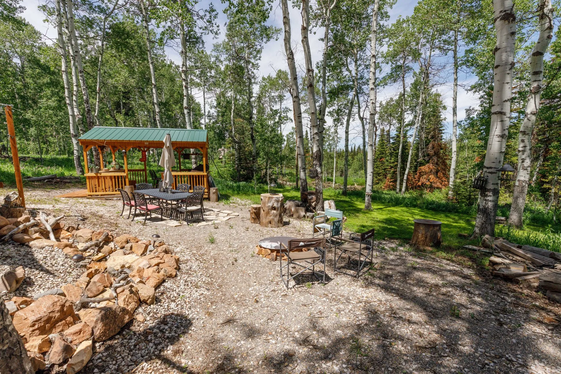 Gravel outdoor area featuring a wood gazebo with a green metal roof, a stone-lined fire pit, and mature trees
