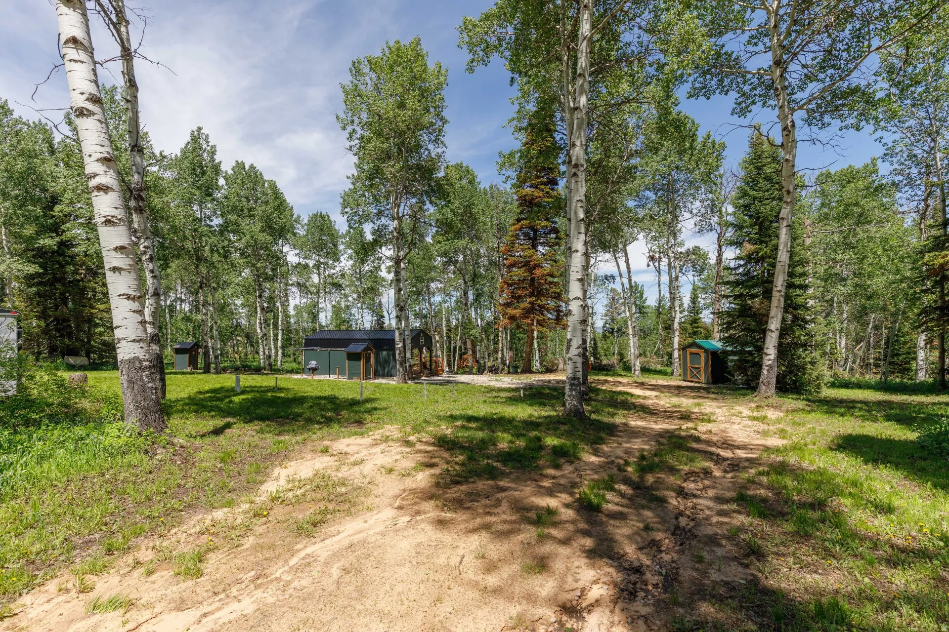 Lush, tree-lined property featuring a green-sided structure with a dark roofline, a smaller wood-sided shed with a green roof, and a separate white-sided structure