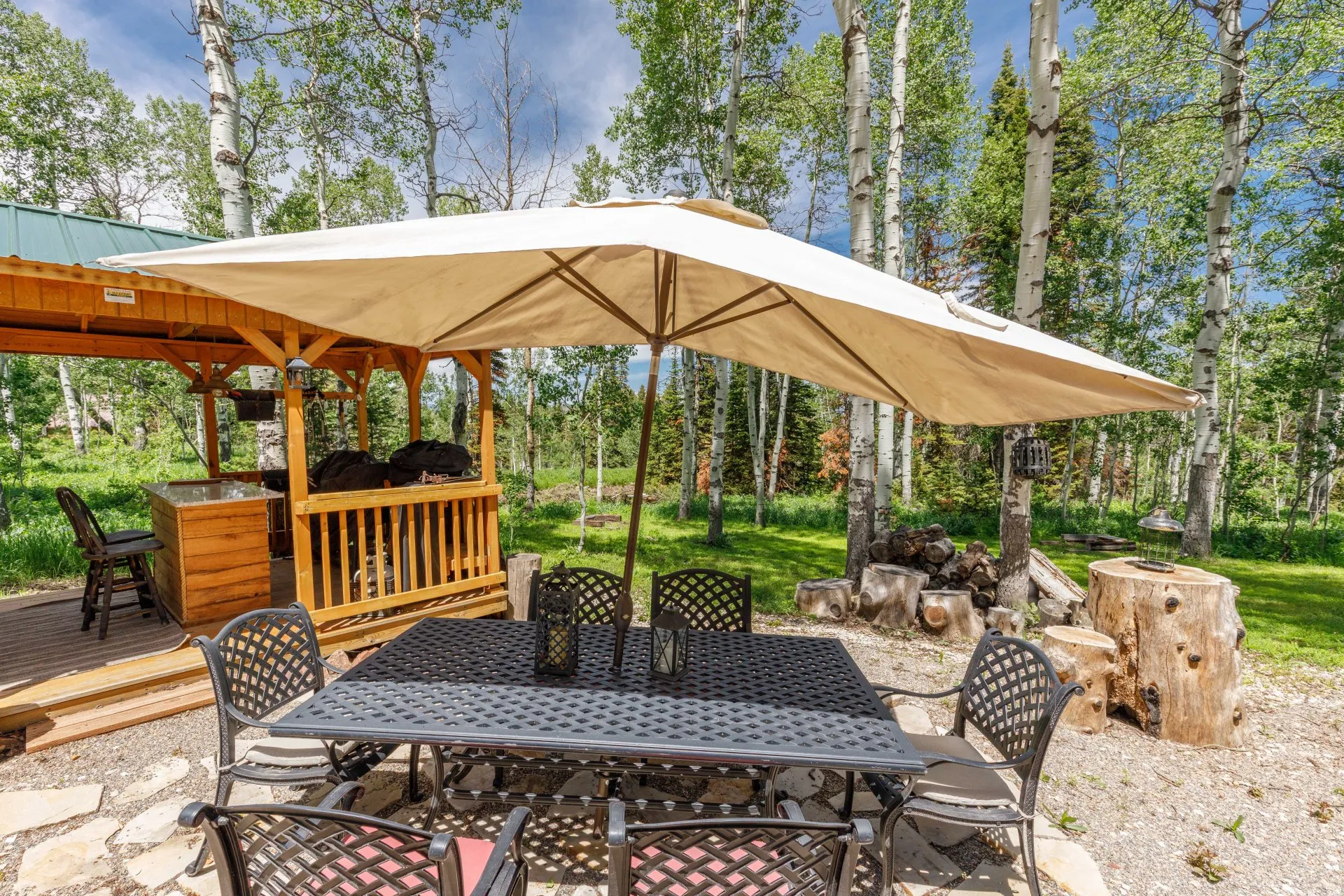 Outdoor dining area featuring a black metal patio set with an umbrella, stone paver patio, and a wooden outdoor kitchen structure with a metal roof