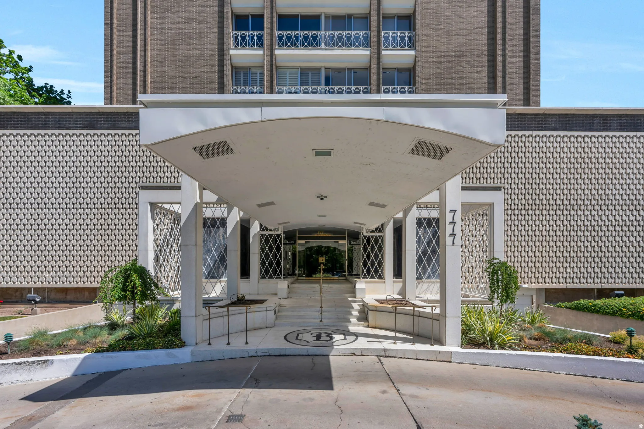 Building entrance featuring a curved portico, patterned facade, integrated planters with landscaping, and a concrete driveway