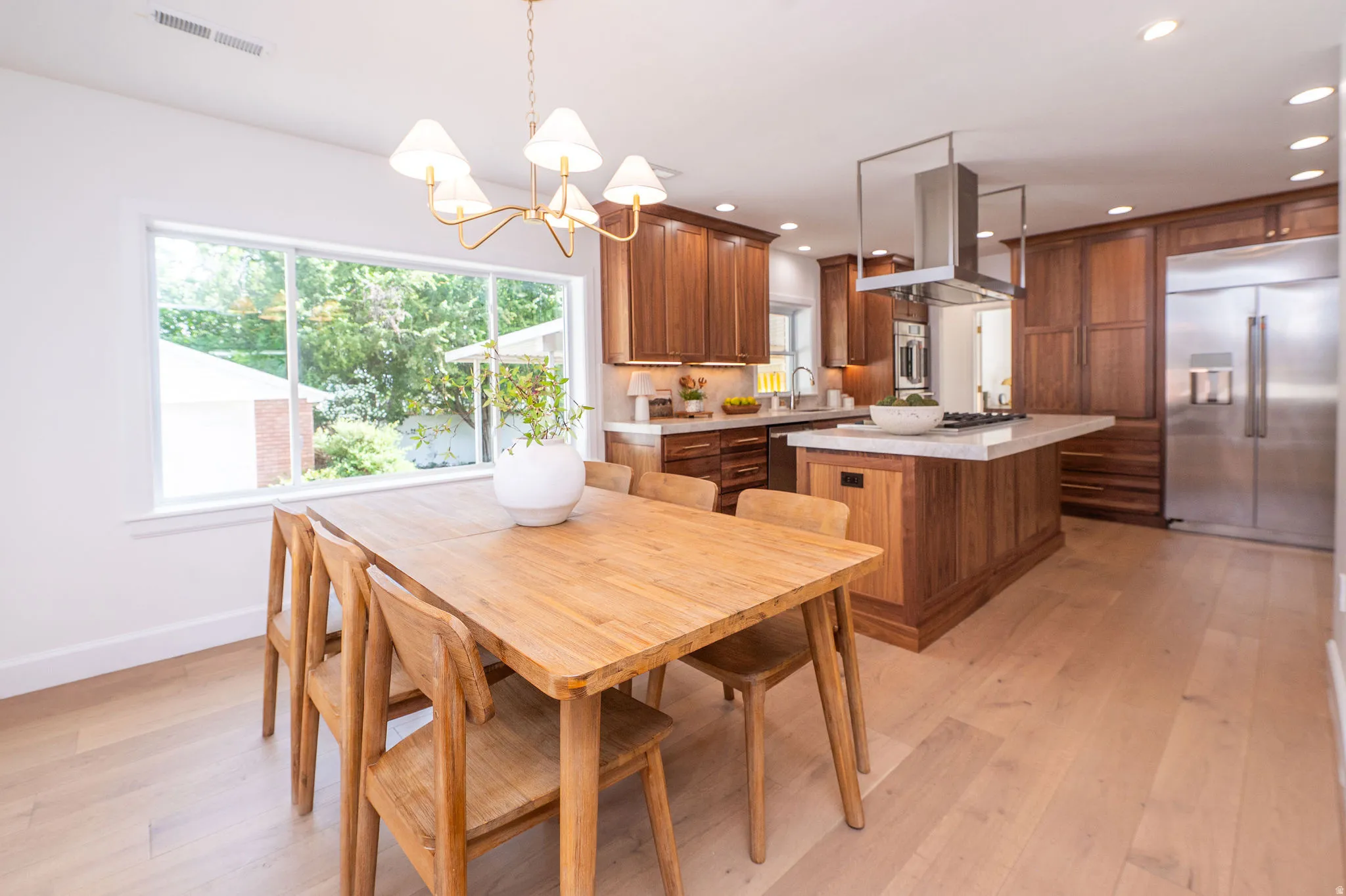 Open-concept kitchen and dining area featuring light wood-finish flooring, extensive wood cabinetry, a large kitchen island with cooktop and overhead exhaust, stainless steel refrigerator, and recessed lighting