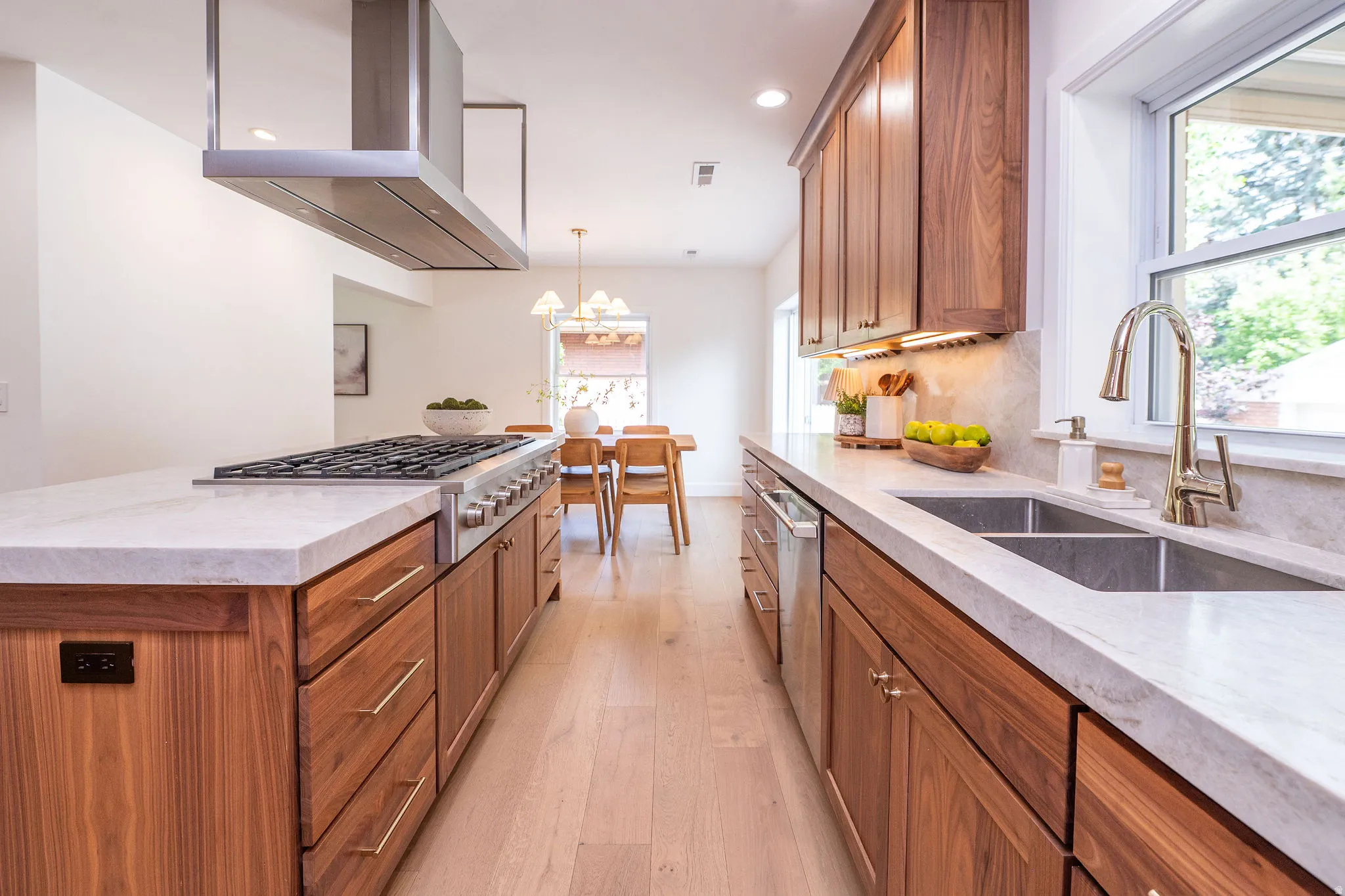 Gourmet kitchen featuring a stainless steel range with overhead hood, wood-finish cabinetry, light-toned countertops, a dual-basin sink, and light wood-finish flooring