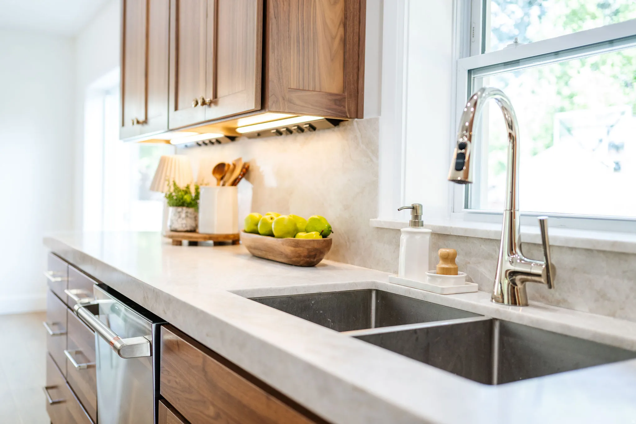 Kitchen featuring a double basin stainless steel sink, a chrome gooseneck faucet, light-toned stone countertops, natural wood-finish cabinetry, and under-cabinet lighting