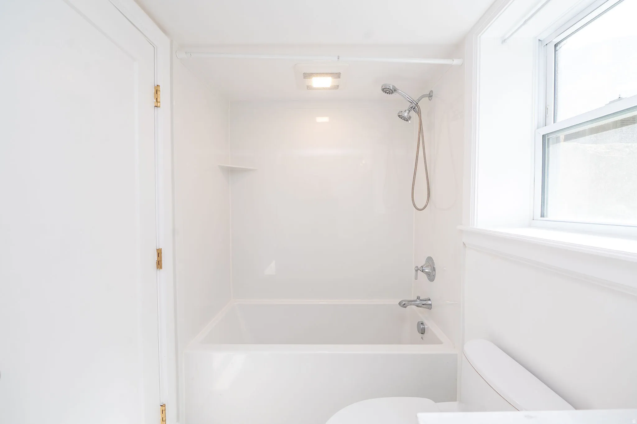 White bathroom featuring a bathtub and shower combination with a handheld showerhead, a toilet, and a window for natural light