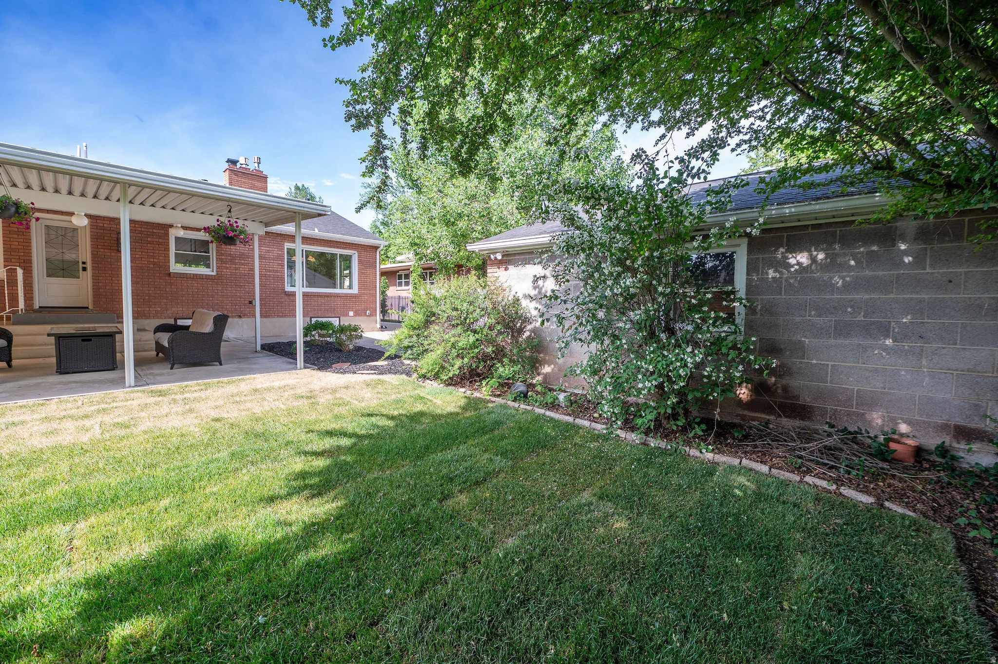 Expansive backyard featuring a covered patio, brick exterior, concrete block wall, and mature trees providing natural shade