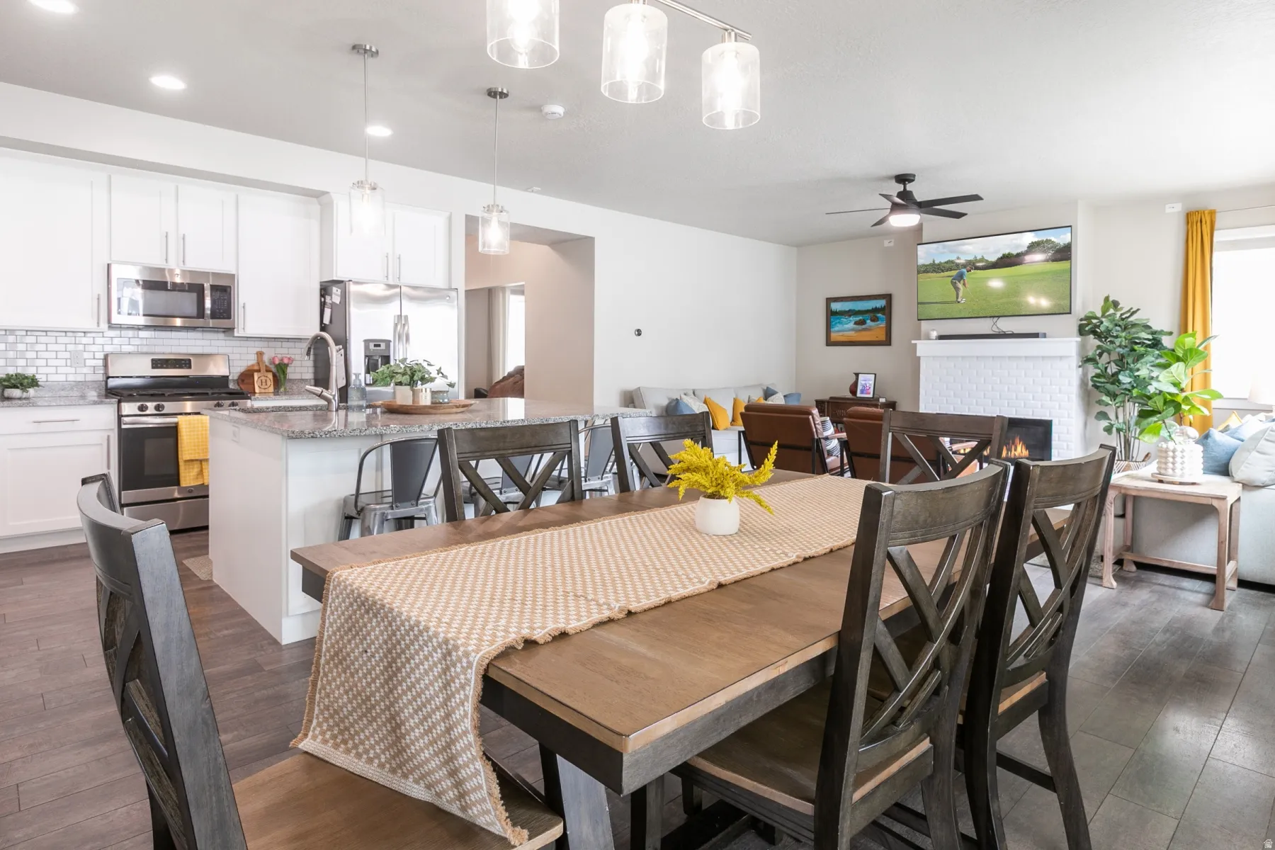 Open-concept living space featuring a dining area with wood-finish flooring, a contemporary kitchen with white cabinetry and stainless steel appliances, and a living room with a white brick fireplace