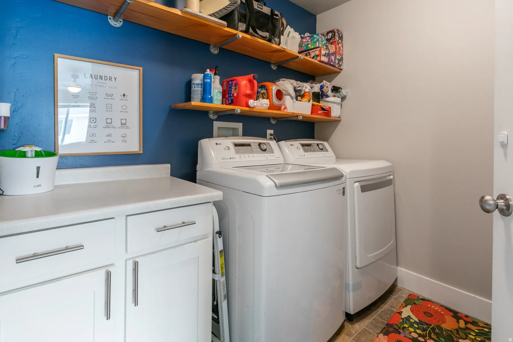 Laundry area featuring white cabinetry, a light-toned countertop, and two wooden floating shelves mounted on a blue accent wall