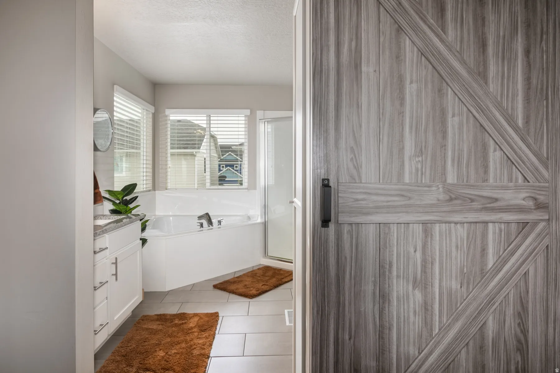 Barn-style wood-finish door with black hardware, opening to a bathroom featuring a corner soaking tub, a stand-up shower, and a vanity with granite-style countertop