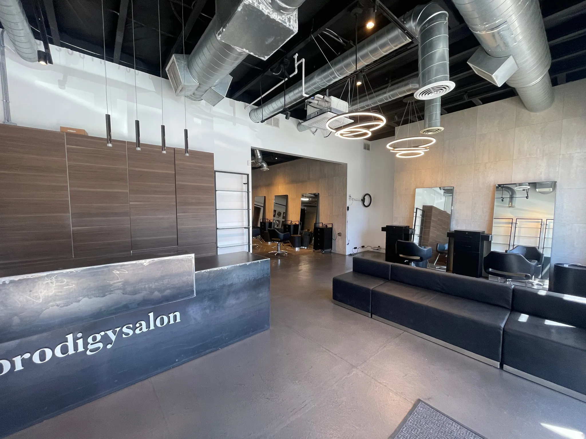 Industrial-style commercial interior featuring exposed ductwork, concrete flooring, modern pendant lighting, a reception desk with wood-finish paneling, and an extended built-in seating area