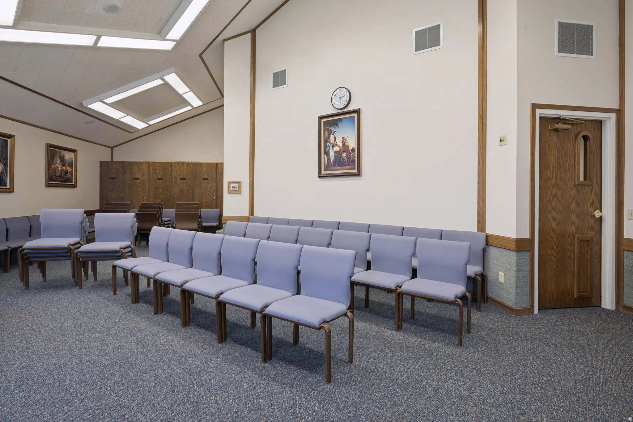 Spacious interior featuring a vaulted ceiling with integrated fluorescent lighting panels, textured wall-to-wall carpeting, wood-paneled accents, and a solid wood entry door with a window.