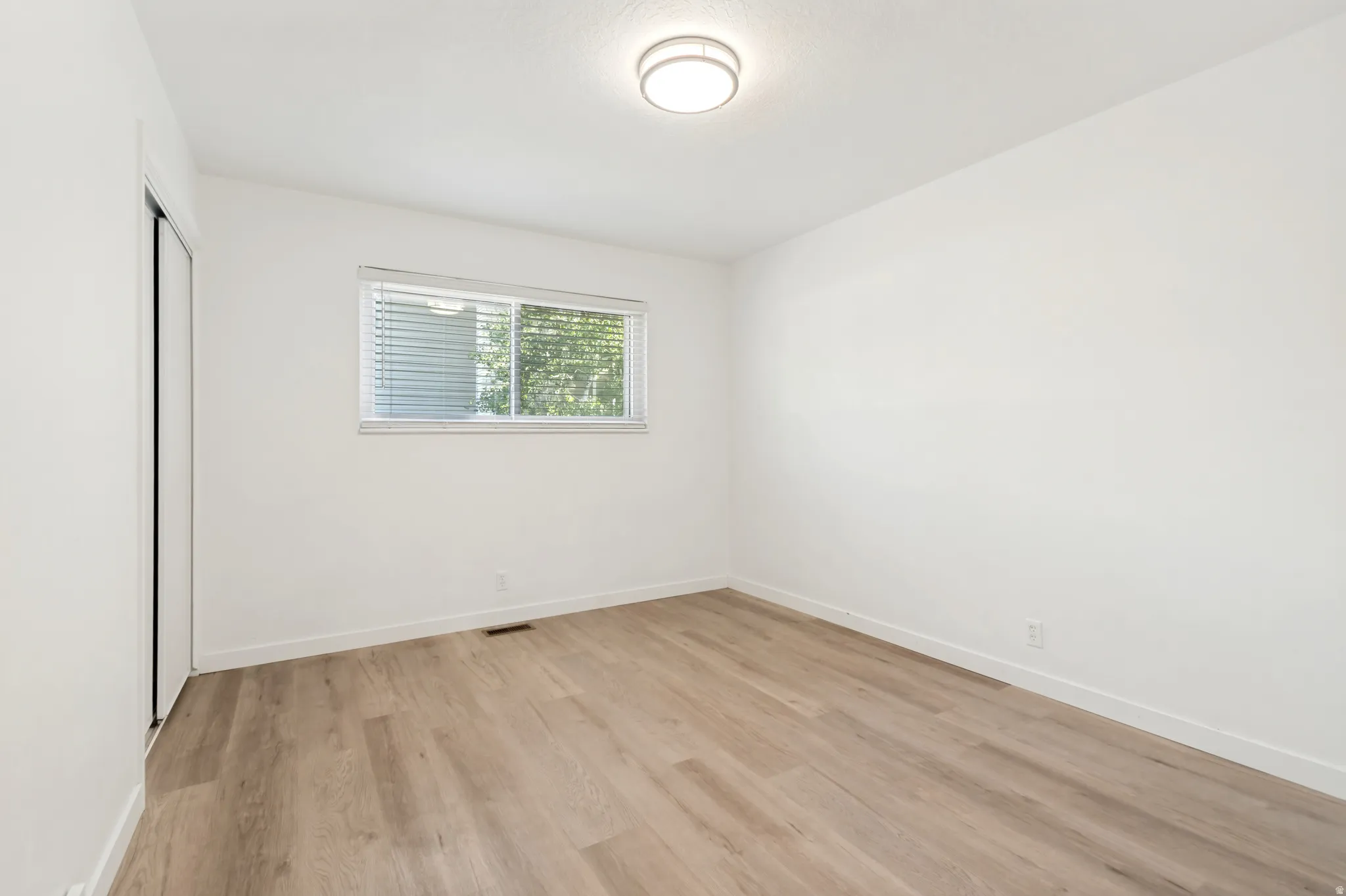 Room featuring wood-finish flooring, a bright window with blinds, and a flush-mount ceiling light