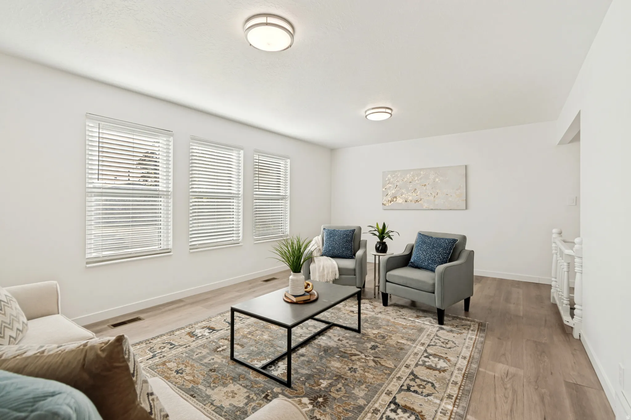 Spacious living area featuring light wood-finish flooring, white wall tones, three large windows, recessed lighting, and a white spindle staircase railing