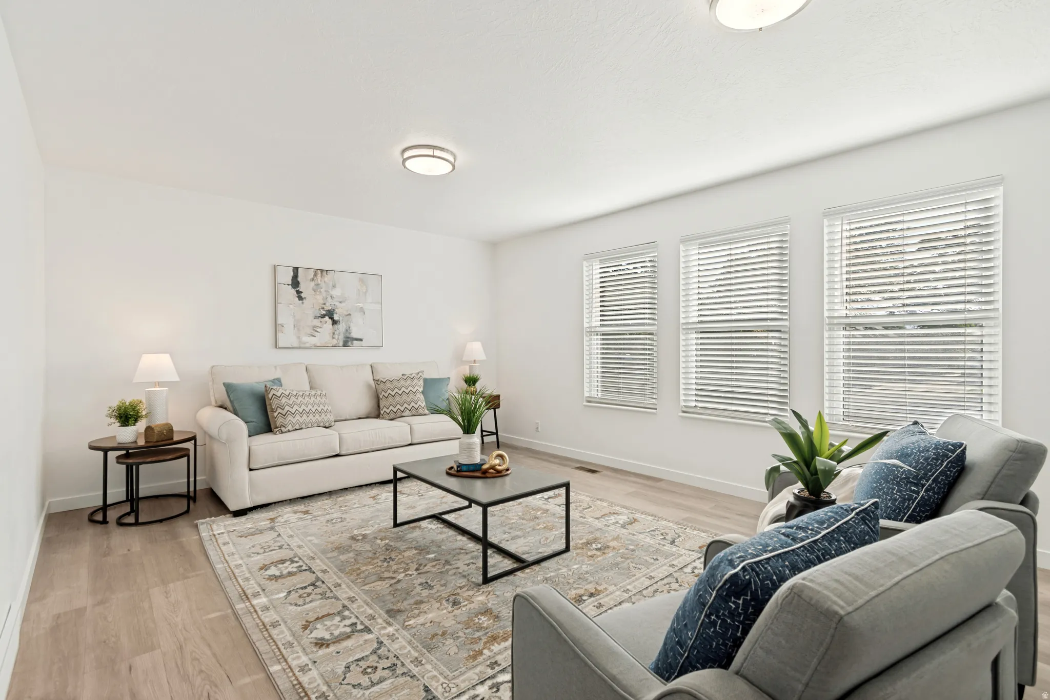 Spacious living area featuring light wood-finish flooring, white walls, and a trio of windows with horizontal blinds