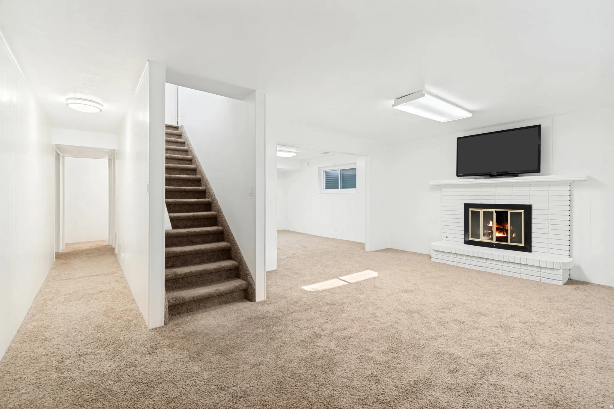 Expansive carpeted room featuring a white brick fireplace with a black insert, a wall-mounted television, and a carpeted staircase