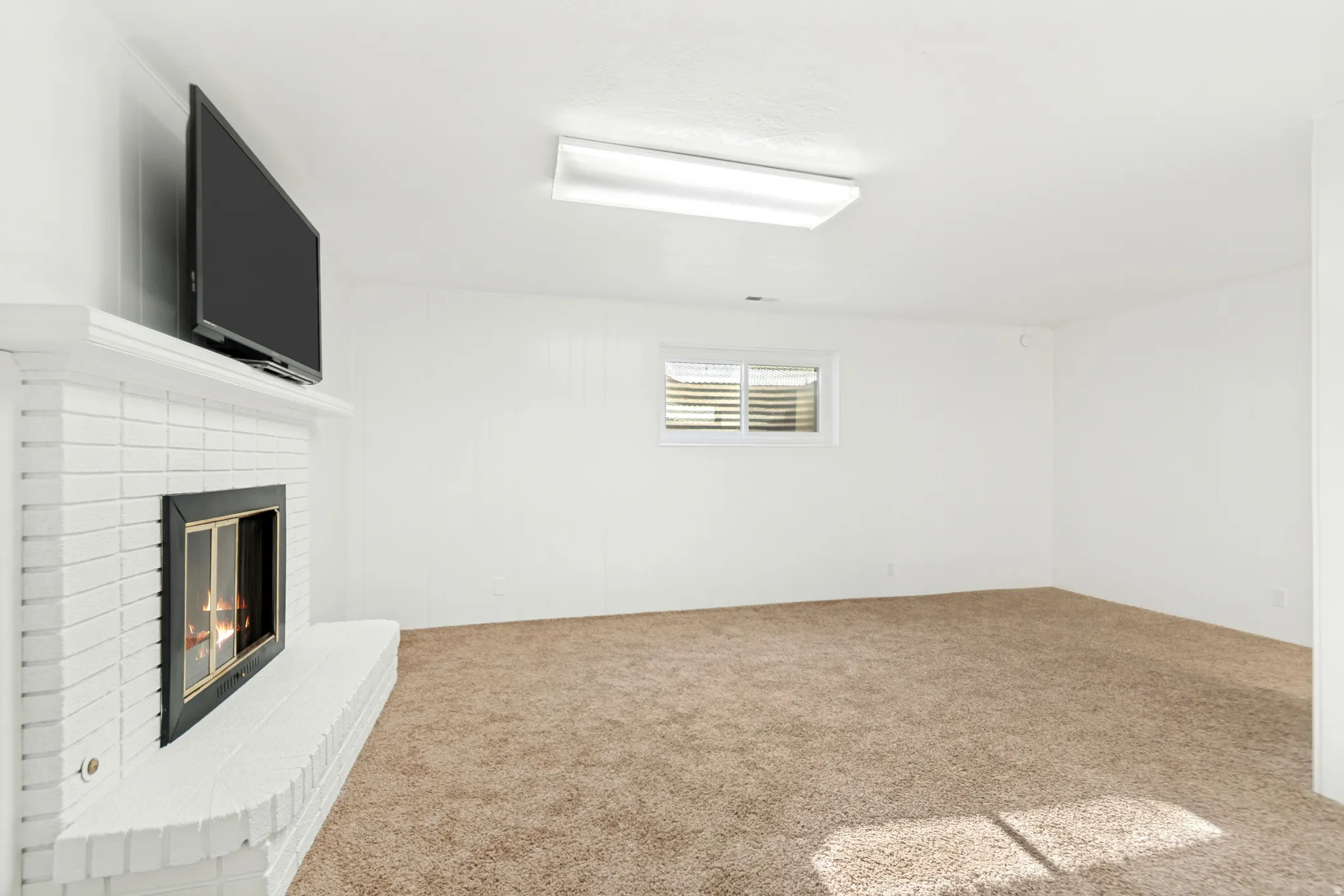 Expansive room featuring a painted brick fireplace with a hearth, wall-to-wall carpeting, a large overhead light fixture, and a window with horizontal blinds