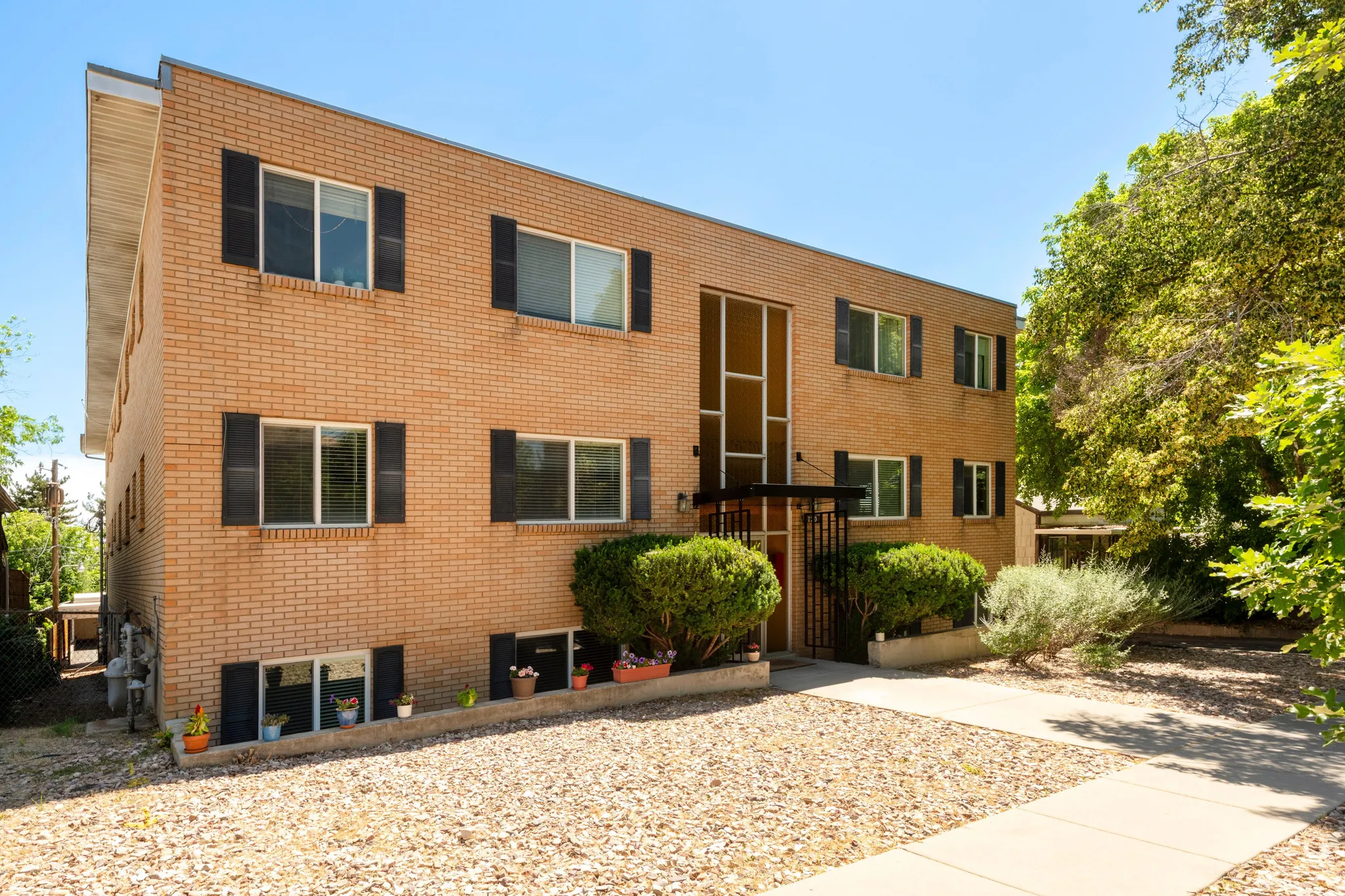 Brick multi-story building featuring multiple windows with dark shutters, a prominent central entrance with an awning, and mature landscaping