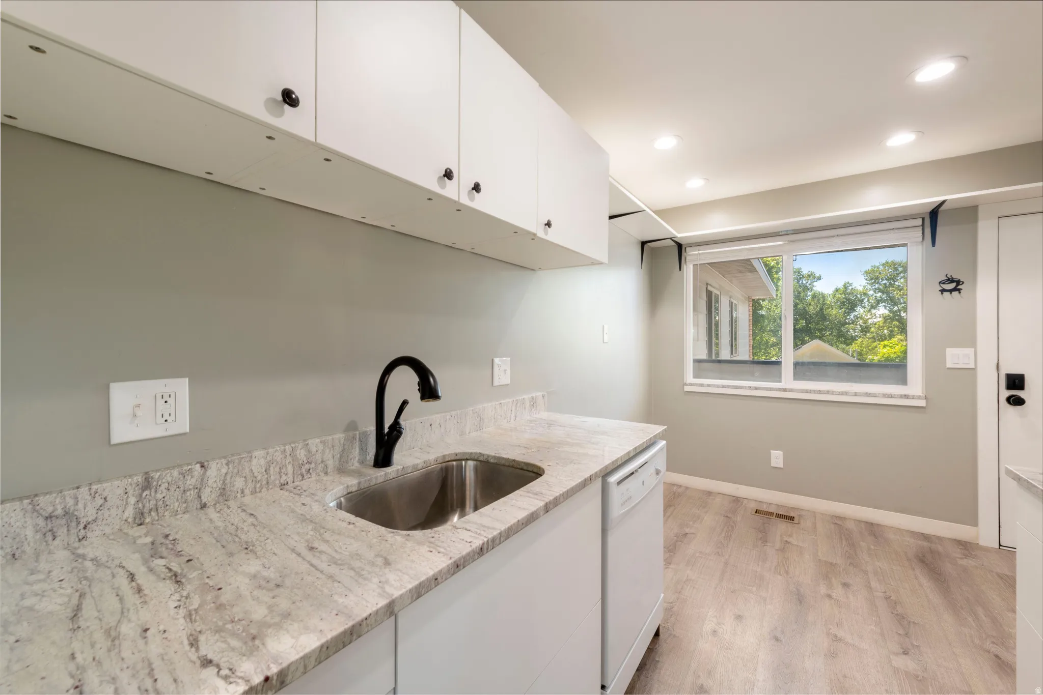 Utility area featuring white upper cabinetry, a stainless steel sink with a matte black faucet, light-toned patterned countertops, and wood-finish flooring