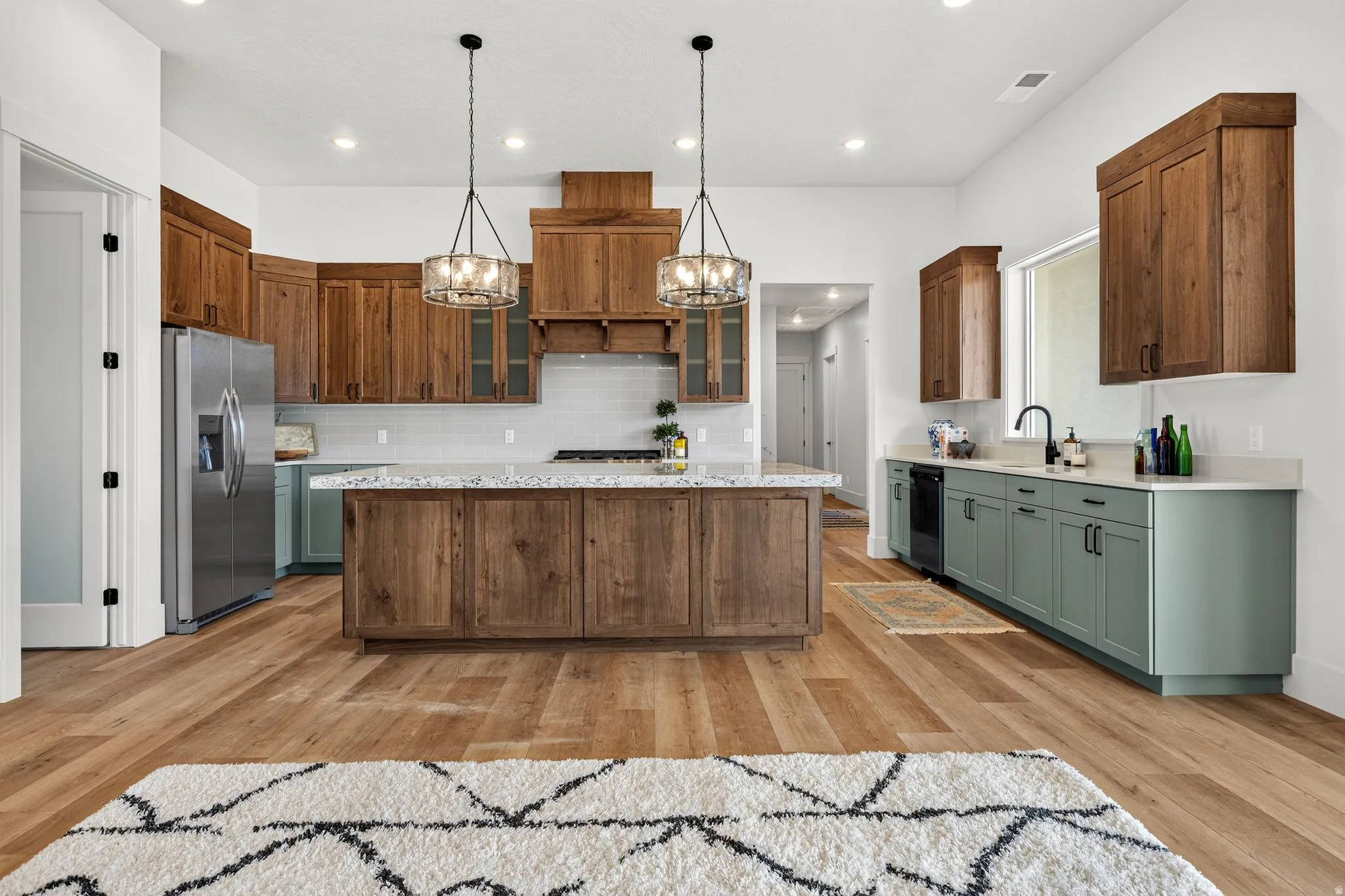 Spacious kitchen featuring a large central island with a light-toned countertop, wood-finish flooring, recessed lighting, stainless steel appliances, and contrasting wood and sage-toned cabinetry