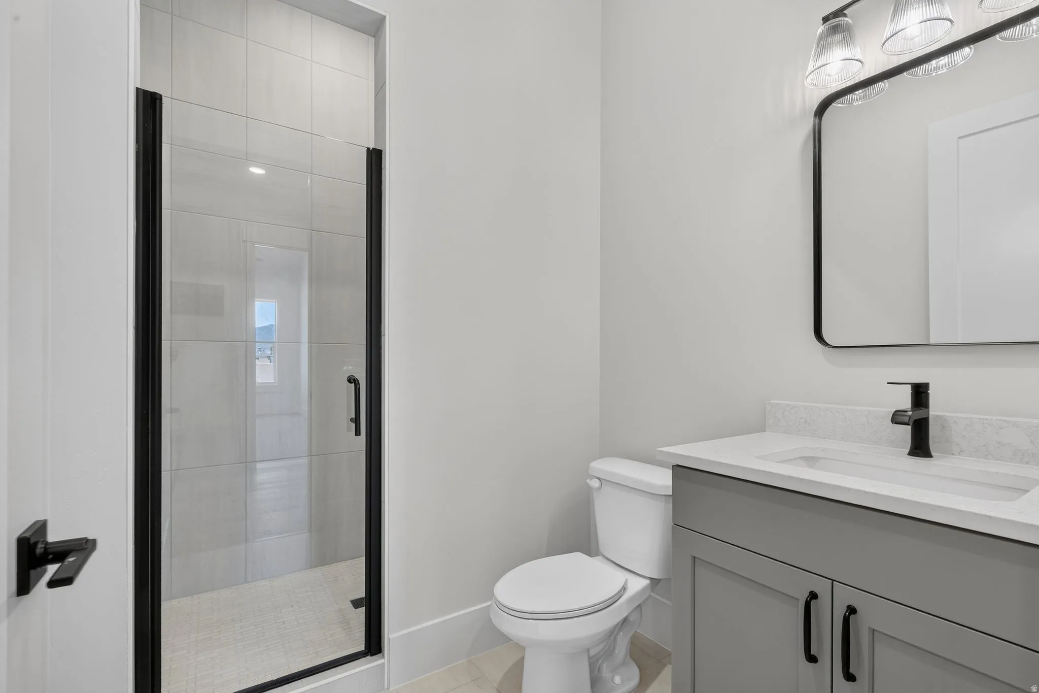 Modern bathroom featuring a walk-in shower with clear glass door and black framing, a light grey vanity with a white countertop and integrated sink, a contemporary black-framed mirror, and a three-light vanity fixture