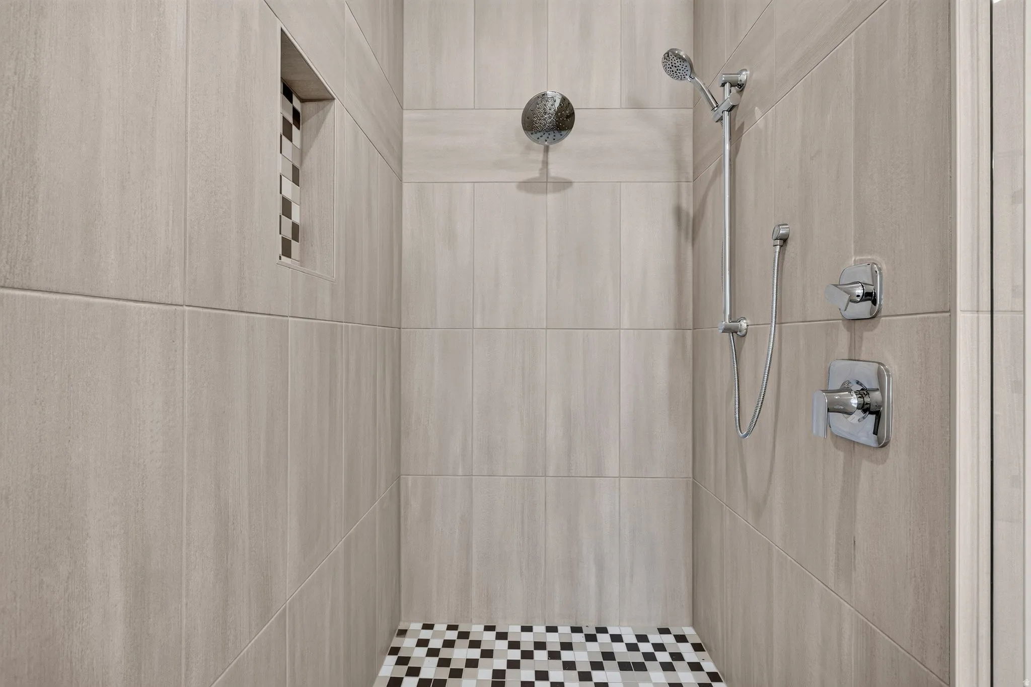 Primary ensuite shower stall featuring light-toned large format wall tile, brown and white mosaic floor tile, a built-in niche with mosaic tile accents, a rain shower head, and a hand-held shower fixture with polished chrome controls