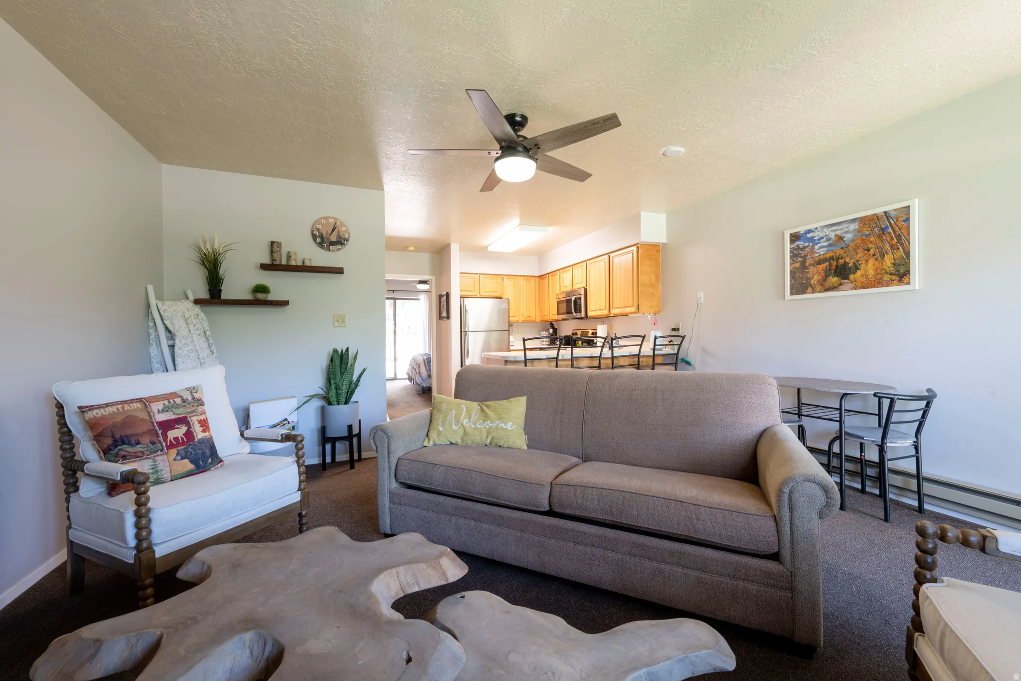 Open-concept living space featuring a ceiling fan, wall-mounted shelving, wood-finish kitchen cabinetry, and an eat-at kitchen counter