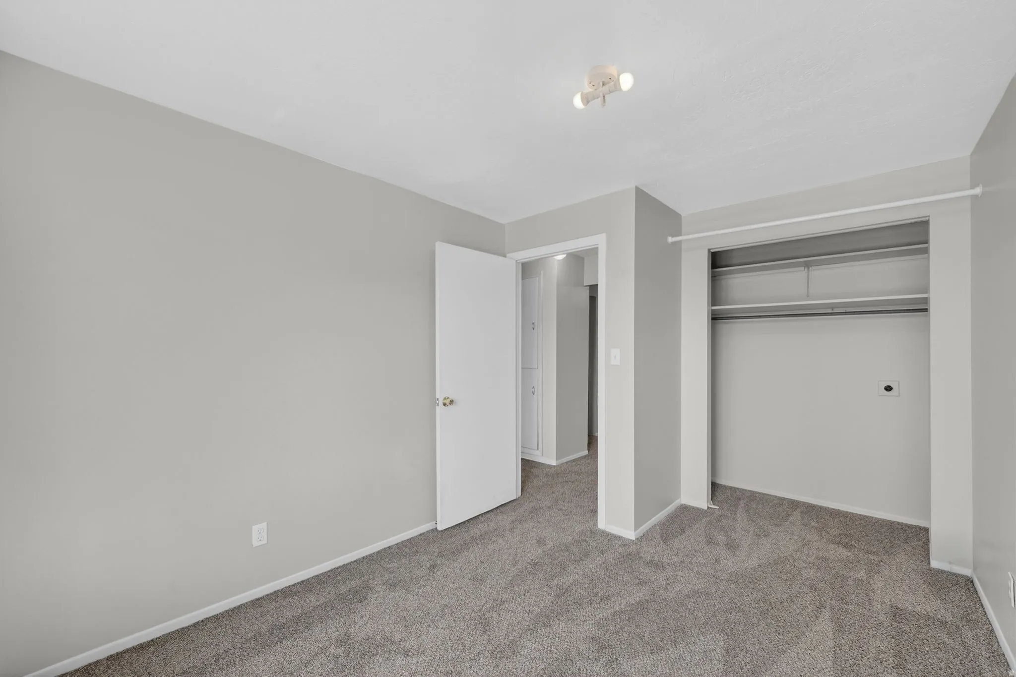 Spacious room featuring gray carpet flooring, light gray wall paint, a built-in closet with shelving and hanging rod, a white interior door, and a ceiling-mounted track light fixture