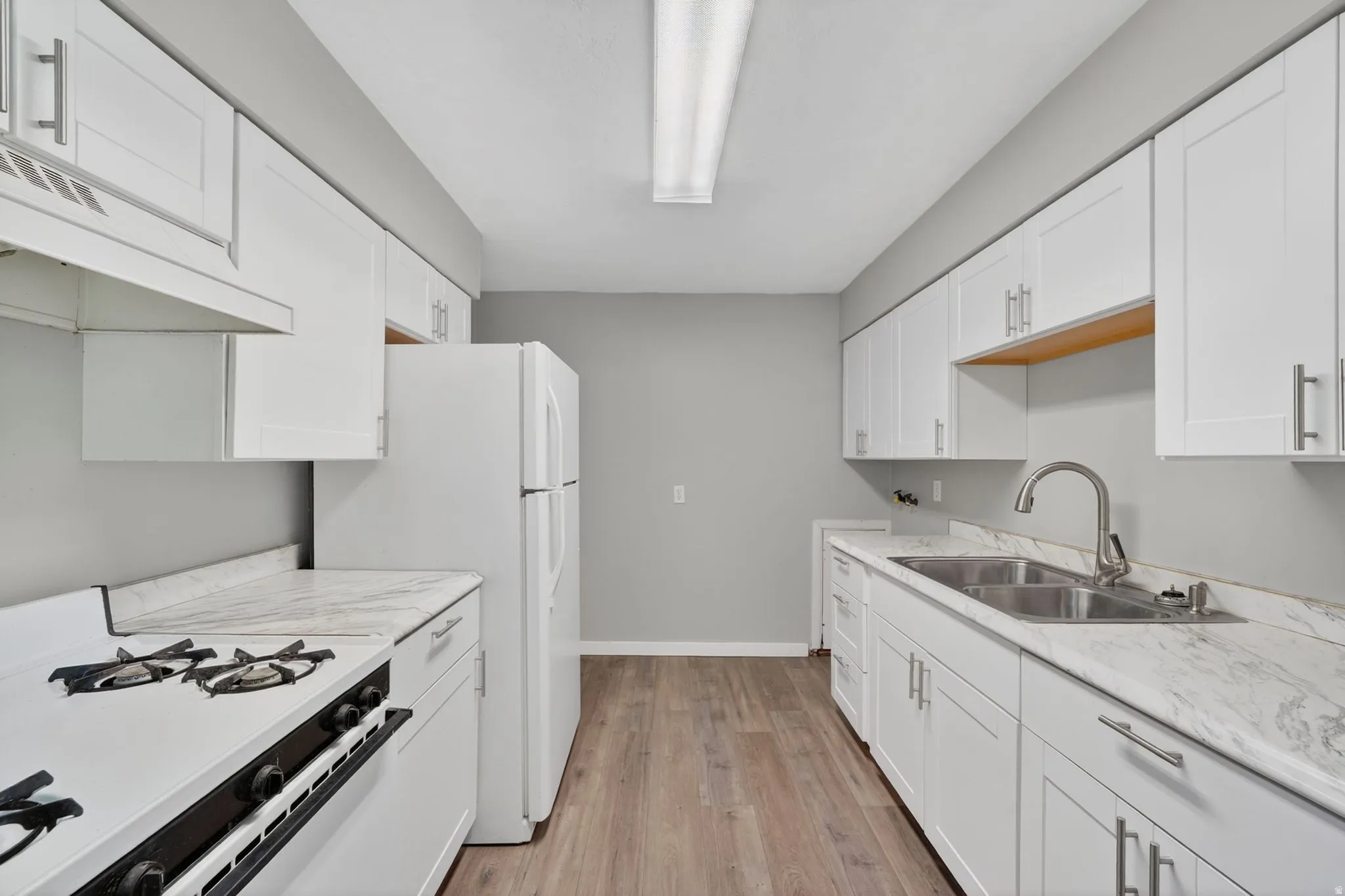 Galley kitchen featuring white cabinetry, wood-finish flooring, a double basin stainless steel sink with a gooseneck faucet, a white gas range, and a white refrigerator