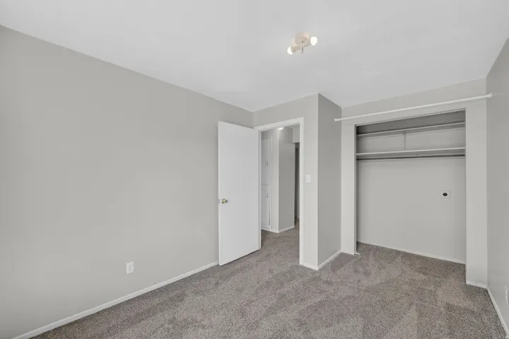Spacious room featuring gray carpet flooring, light gray wall paint, a built-in closet with shelving and hanging rod, a white interior door, and a ceiling-mounted track light fixture