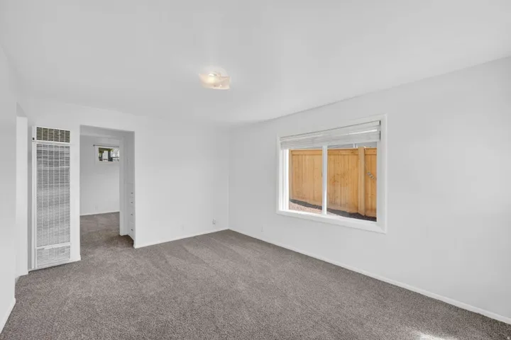 Spacious room with plush gray carpeting, white walls, a large window with privacy blinds, and a wall-mounted heating unit