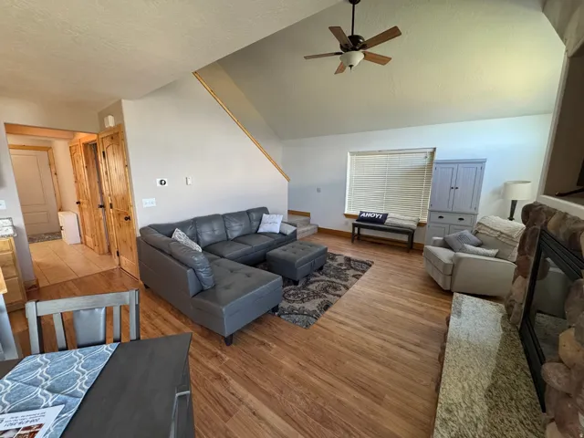Vaulted ceiling living area featuring wood-finish flooring, a stone-surround fireplace with a granite-style hearth, and a ceiling fan