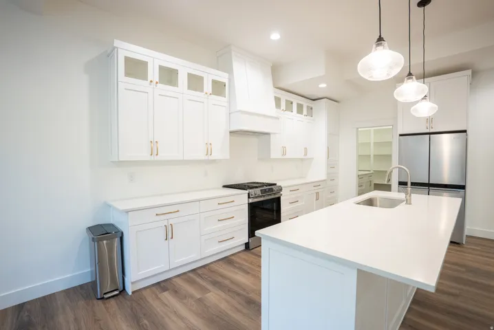 Bright kitchen featuring an island with an undermount sink, white cabinetry with gold-tone hardware, wood-finish flooring, stainless steel appliances, and pendant lighting