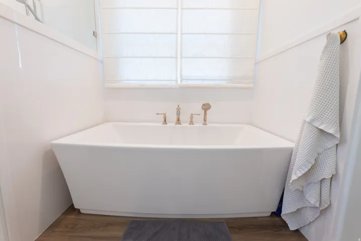 Freestanding soaking tub with gold-tone fixtures, wood-finish flooring, and white paneled walls