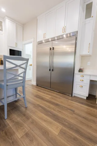 Gourmet kitchen featuring a built-in stainless steel refrigerator, extensive white cabinetry with gold-toned hardware, wood-finish flooring, and a built-in desk area with quartz-style countertops