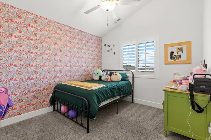 Vaulted ceiling bedroom featuring a window with plantation shutters, a patterned accent wall, plush carpet flooring, and a ceiling fan